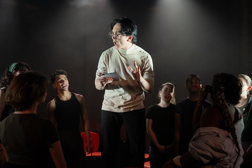 Action shot of the spring 2024 Shakespeare in Performance at RADA final presentation of Julius Caesar. An actor wearing glasses and holding a sheet of paper delivers a monologue while other actors surround him in mid-performance.